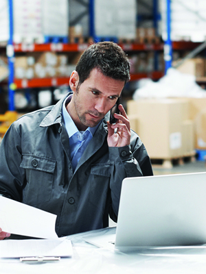 A guy in a warehouse talking on the phone while looking at his computer. 